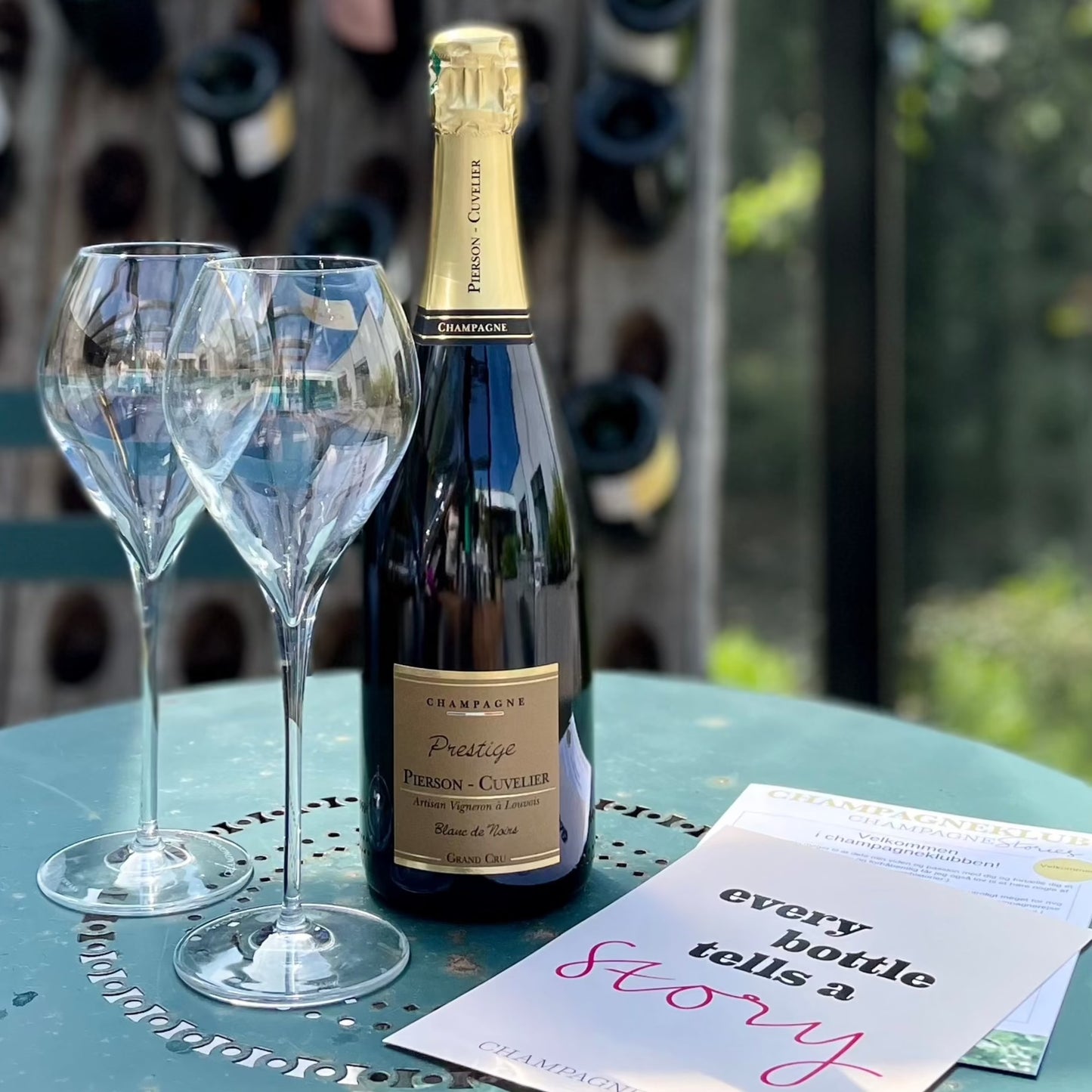 På bord står 2 champagneglas 41 cl, 1 flaske champagne Pierson-Cuvelier Prestige. Ved siden af ligger 1 klubflyer og 1 postkort med teksten "every bottle tells a stories - Champagne Stories"