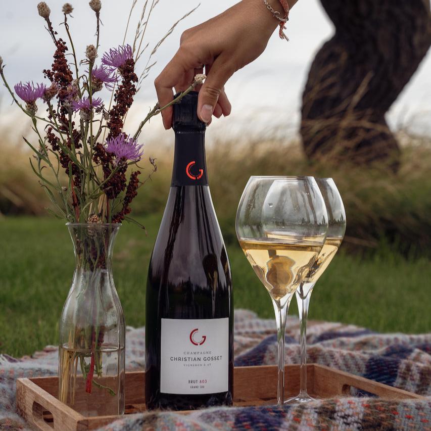 Champagneflaske på bakke med 2 fyldte gls og en buket markblomster i vase, på et picnictæppe på græsplæne, kvindehånd holder om proppen på flasken, træ i baggrund, Champagne Christian Gosset, Brut A03, Grand Cru