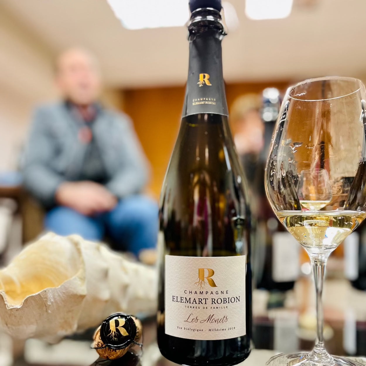 Champagne flaske på glasbord med konkylie og et champagneglas ved siden, champagneprop med Logo R, uskarpt i baggrunden ses Thierry Robion, Champagne Elemart Robion, Les Monets, Millésime 2018, økologisk champagne, årgangschampgne