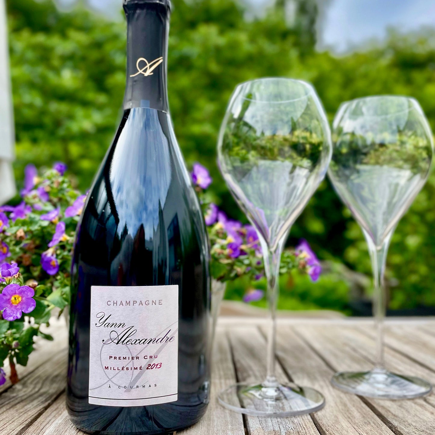 Champagne flaske på havebord med lilla små blomster og grøn baggrund, 2 tomme champagneglas står ved siden af, hvid etikette, Champagne Yann Alexandre, Premier Cru, Millésimé 2013