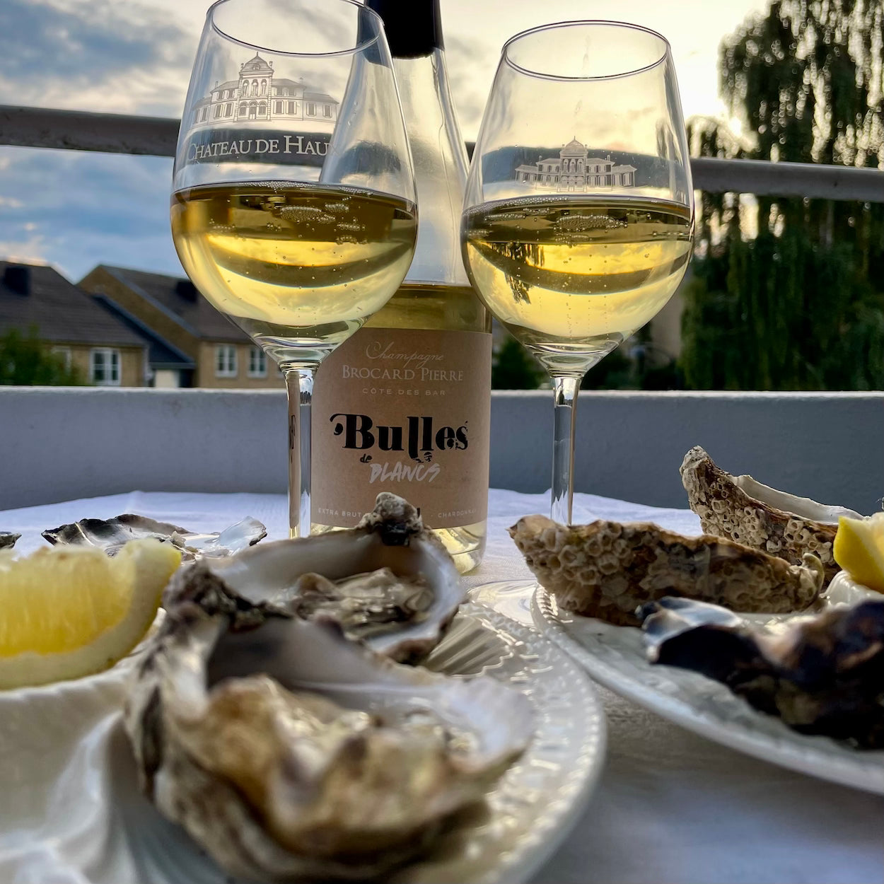 2 champagneglas fyldt med champagne og champagne flaske i baggrund, himmel og huse, østers og citron i forgrunden, Champagneflaske på hvid baggrund, Champagne Brocard Pierre, Bulles de Blancs