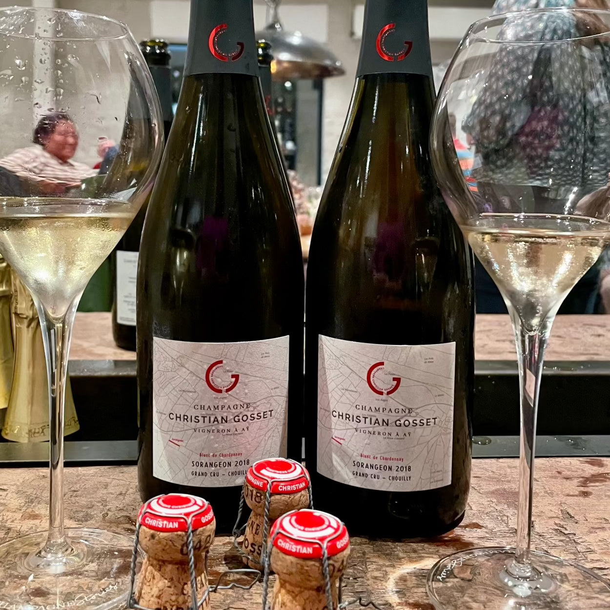 2 flaske champagne på bord med 2 champagneglas og 3 champagnepropper, mennesker i baggrunden, Champagneflaske på bord med champagneglas foran fyldt  med rosé champagne, Champagne Christian Gosset, Sorangeon 2018, Blanc de Chardonnay, Grand Cru