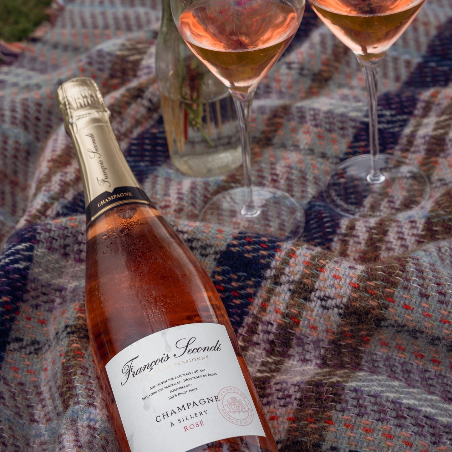 Champagne flaske, Francois Seconde champagne, Rosé champagne, brut, Flasken ligger på et ternet picnic tæppe og 2 glas med rosé champagne står på tæppet