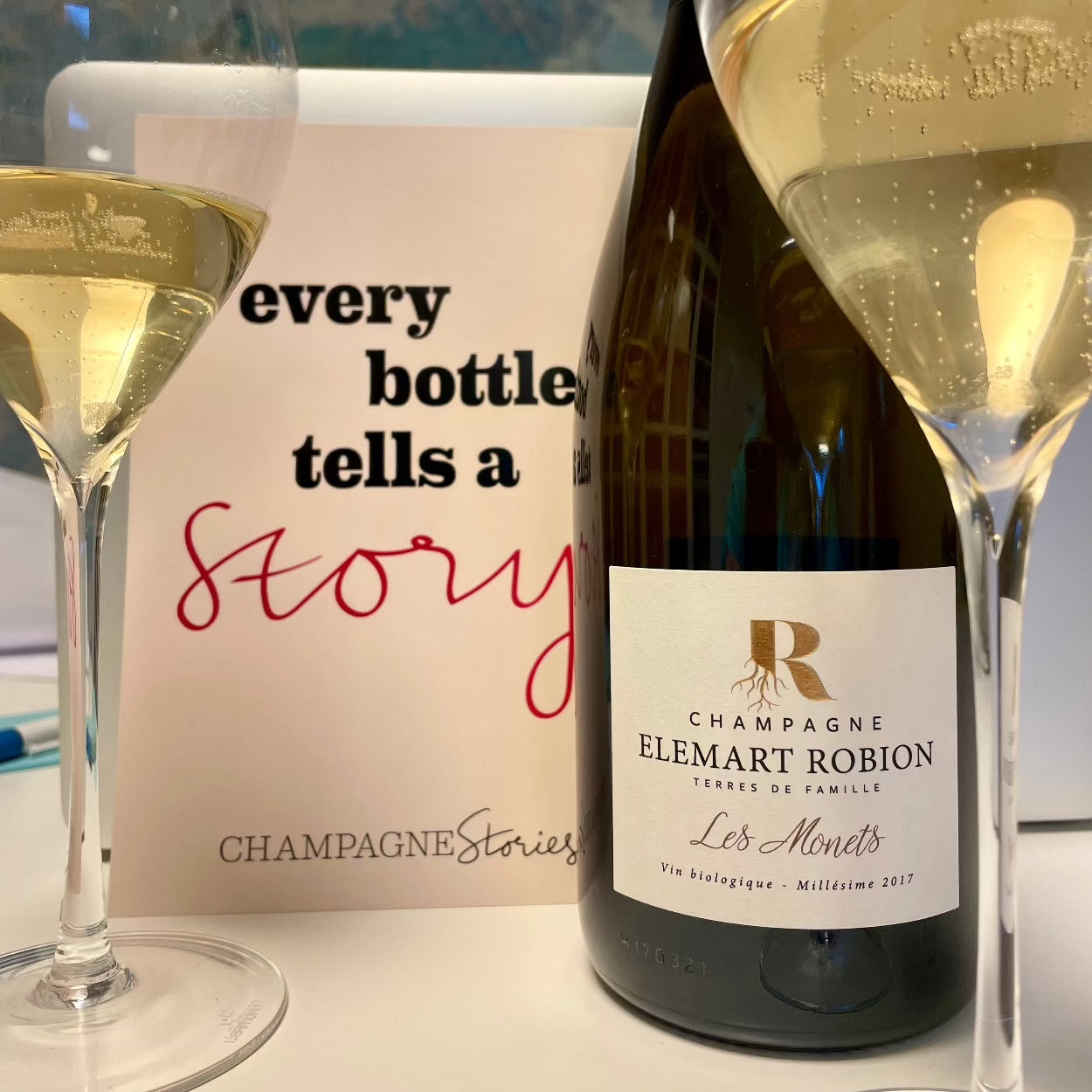 Champagne flaske på bord med 2 champagneglas fyldt med champagne og et postkort med teksten "every bottle tells a story", bobler i glasset, Champagne Elemart Robion, Les Monets, Millésime 2018, økologisk champagne, årgangschampgne