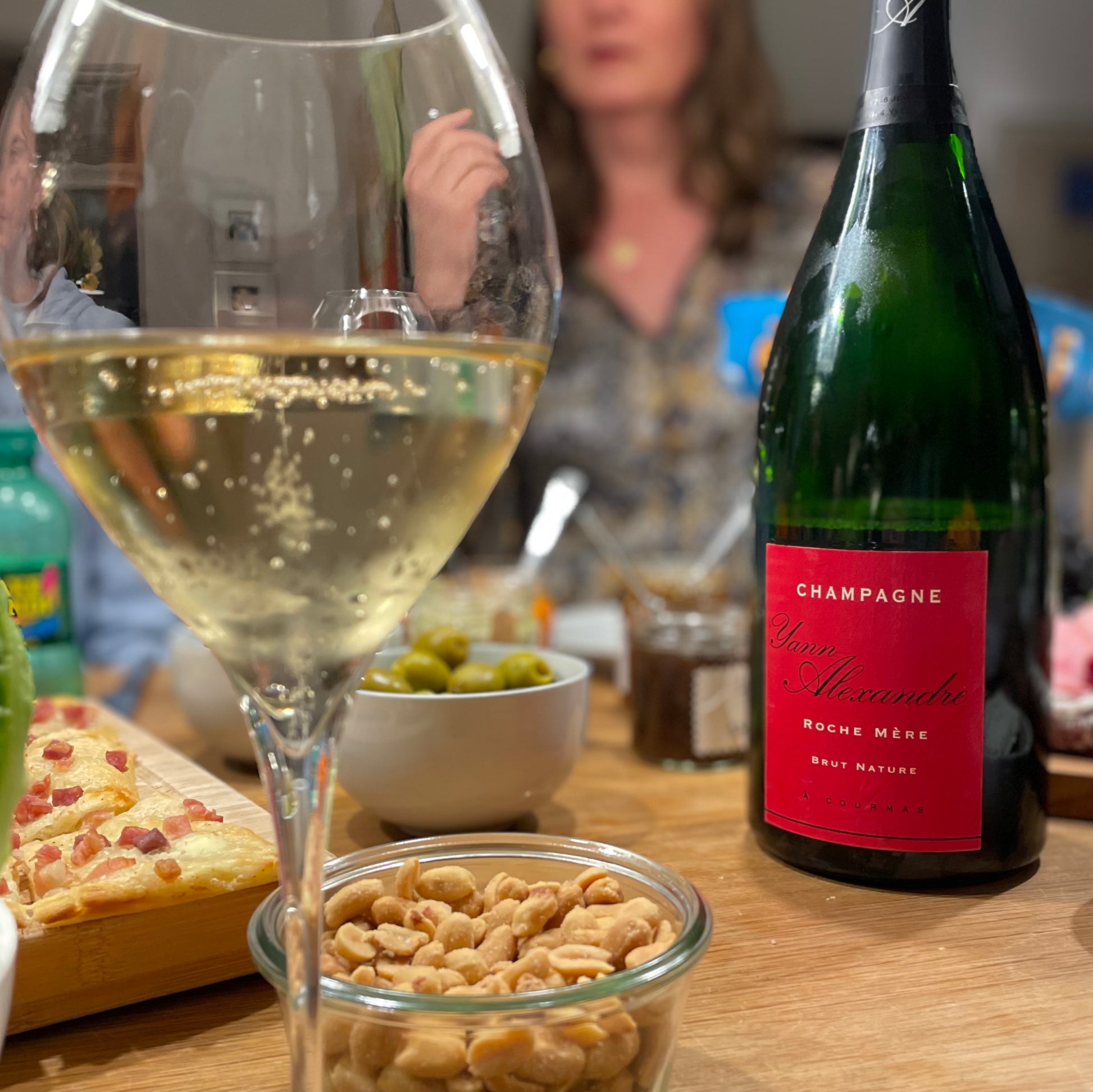 Champagne flaske på bord med tapas, peanuts, oliven, uskarp personer i baggrund, tæt på champagneglas med champagne i forgrunden, bobler i glasset, rød etikette, Champagne Yann Alexandre, Roche Mere, Brut Nature