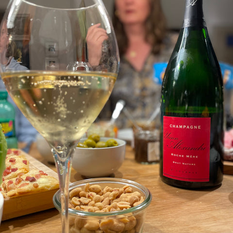 Champagne flaske på bord med tapas, peanuts, oliven, uskarp personer i baggrund, tæt på champagneglas med champagne i forgrunden, bobler i glasset, rød etikette, Champagne Yann Alexandre, Roche Mere, Brut Nature