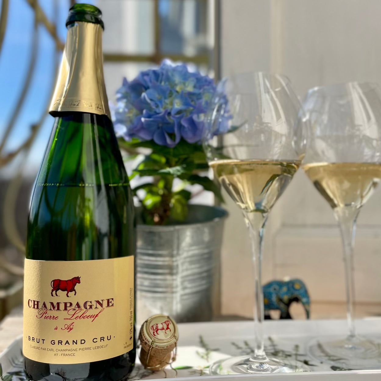 1 flaske champagne, 2 glas med champagne og en blomst i baggrunden, champagneprop ved flasken, Pierre Leboeuf champagne, Brut Grand Cru, gul etikette med rød okse og rød skrift