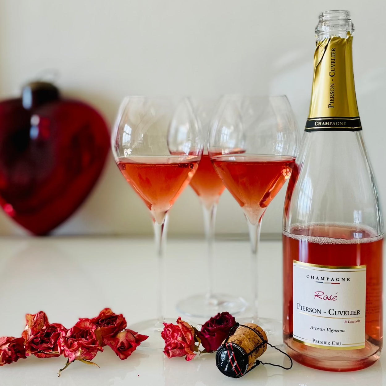 1 flaske rosé champagne fra Champagne Pierson-Cuvelier, Rosé, Premier Cru, ved siden af 3 Lehmann Grand Jamesse champagneglas med rosé champagne, rødt hjerte i baggrund, forest tørret rosenknopper og en champagnepror, hvid baggrund