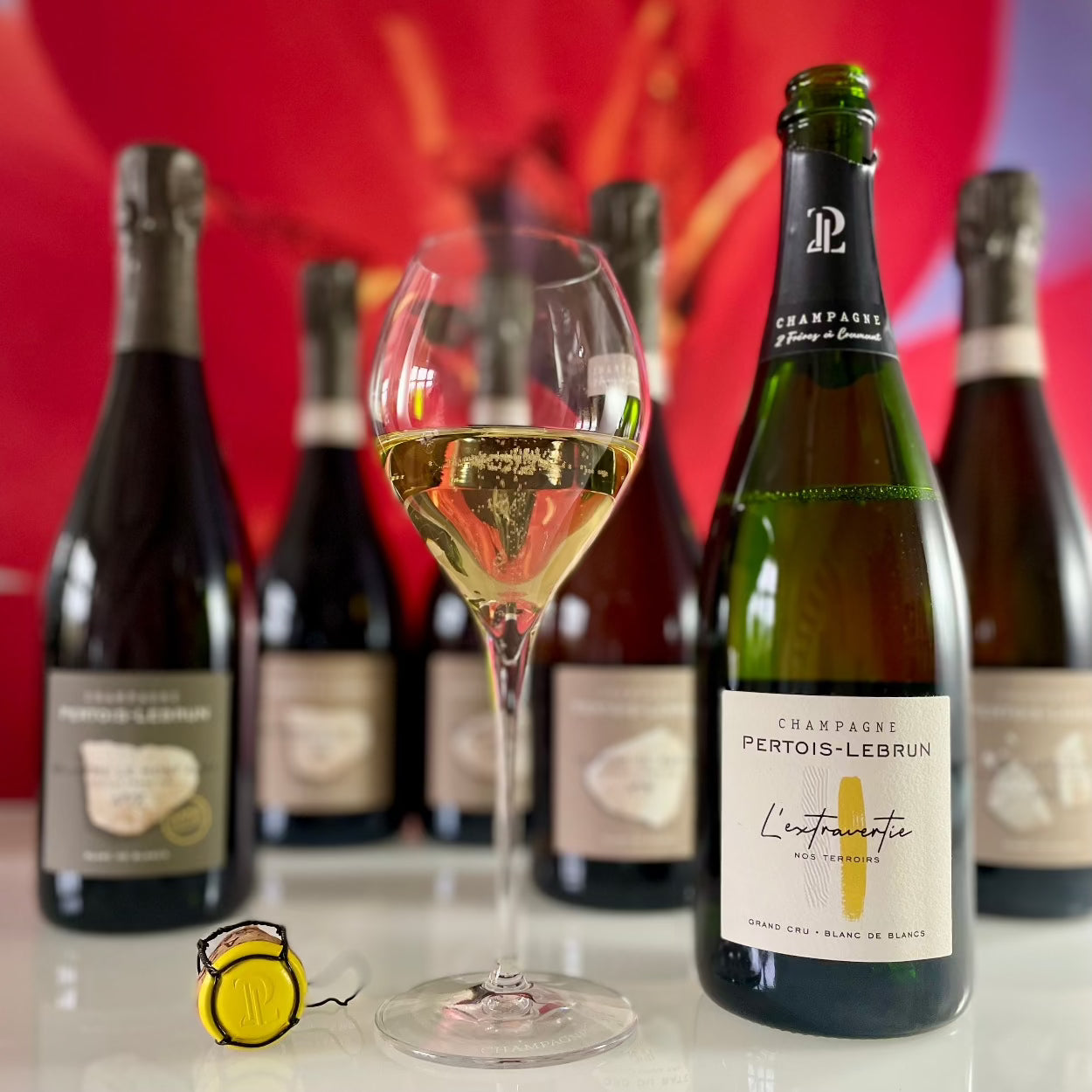 Flere flasker champagne på bord med rød baggrund, champagneprop med gul kapsel og logo "PL", Champagne Pertois-Lebrun, L'extravertie, Grand Cru, Blanc de Blancs