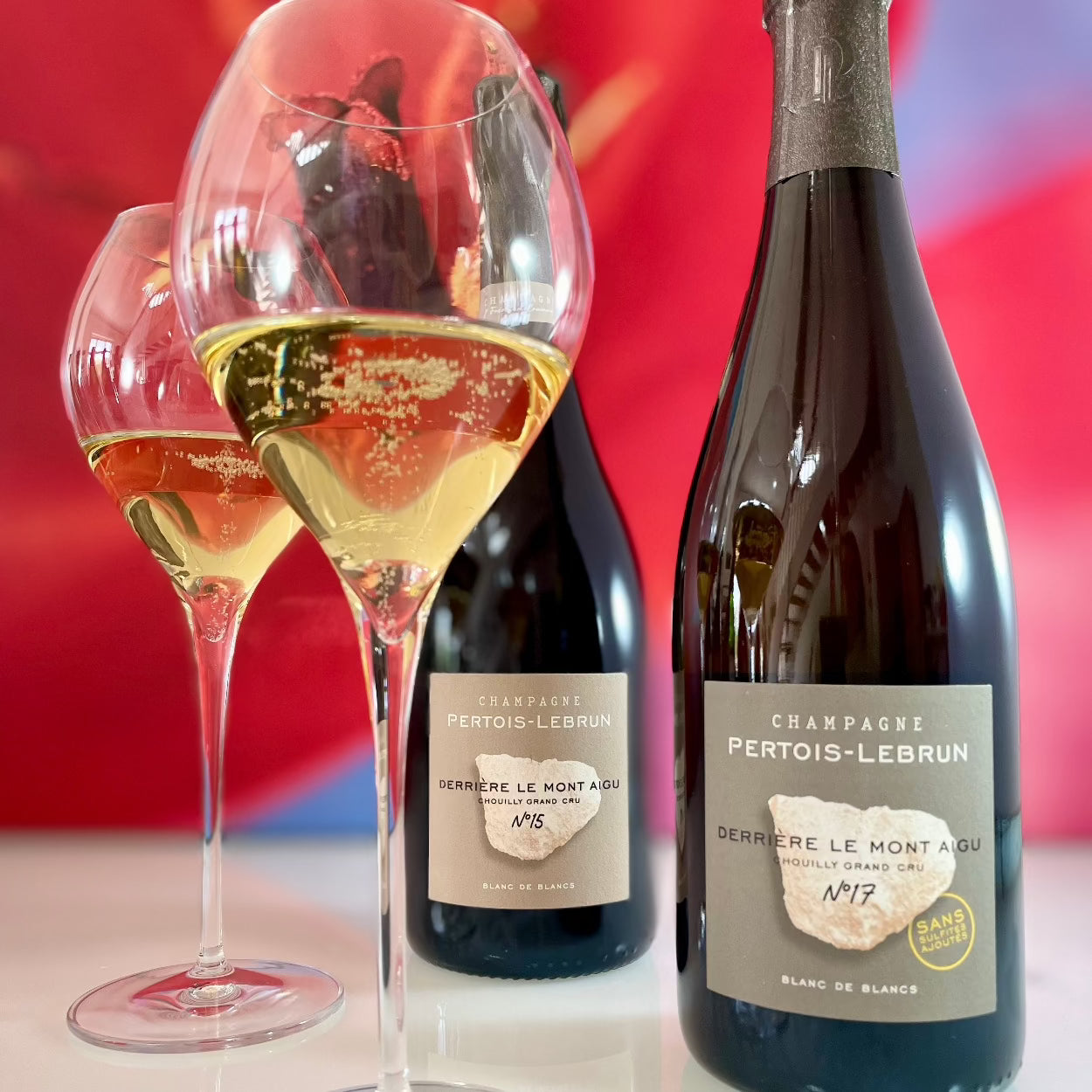 2 champagneglas med champagne og 2 champagneflasker, rød uskarp baggrund, Champagne Pertois-Lebrun, Derriére le Mont Aigu no. 15, Derriére le Mont Aigu no. 17, Grand Cru, årgangschampagne