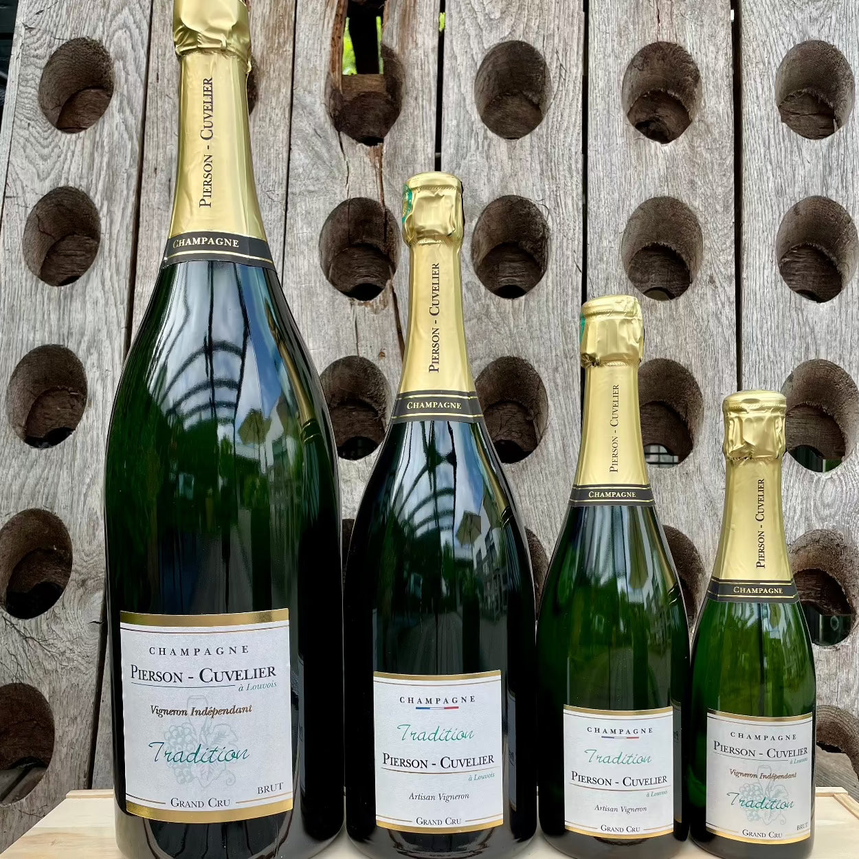 4 flasker champagne fra Champagne Pierson-Cuvelier, Tradition, Grand Cru, 4 forskellige størrelser flaske, 1/2 flaske 0,37 liter, normal flaske 0,75 liter, magnum flaske 1,5 liter, jerobaum 3 liter, baggrund er en pupitre, træ plade med huller til champagneflasker