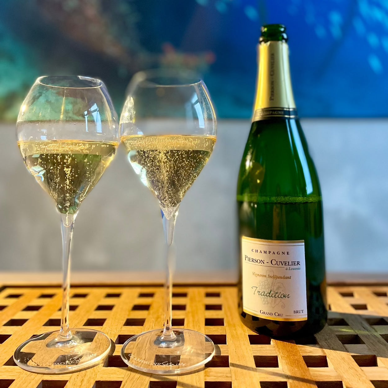 1 flaske champagne fra Champagne Pierson-Cuvelier, Tradition, Grand Cru, til venstre for flasken står 2 Lehmann Grand Jamesse champagneglas fyldt med champagne, bobler i glasset, står på en træbakke, uskarp baggrund