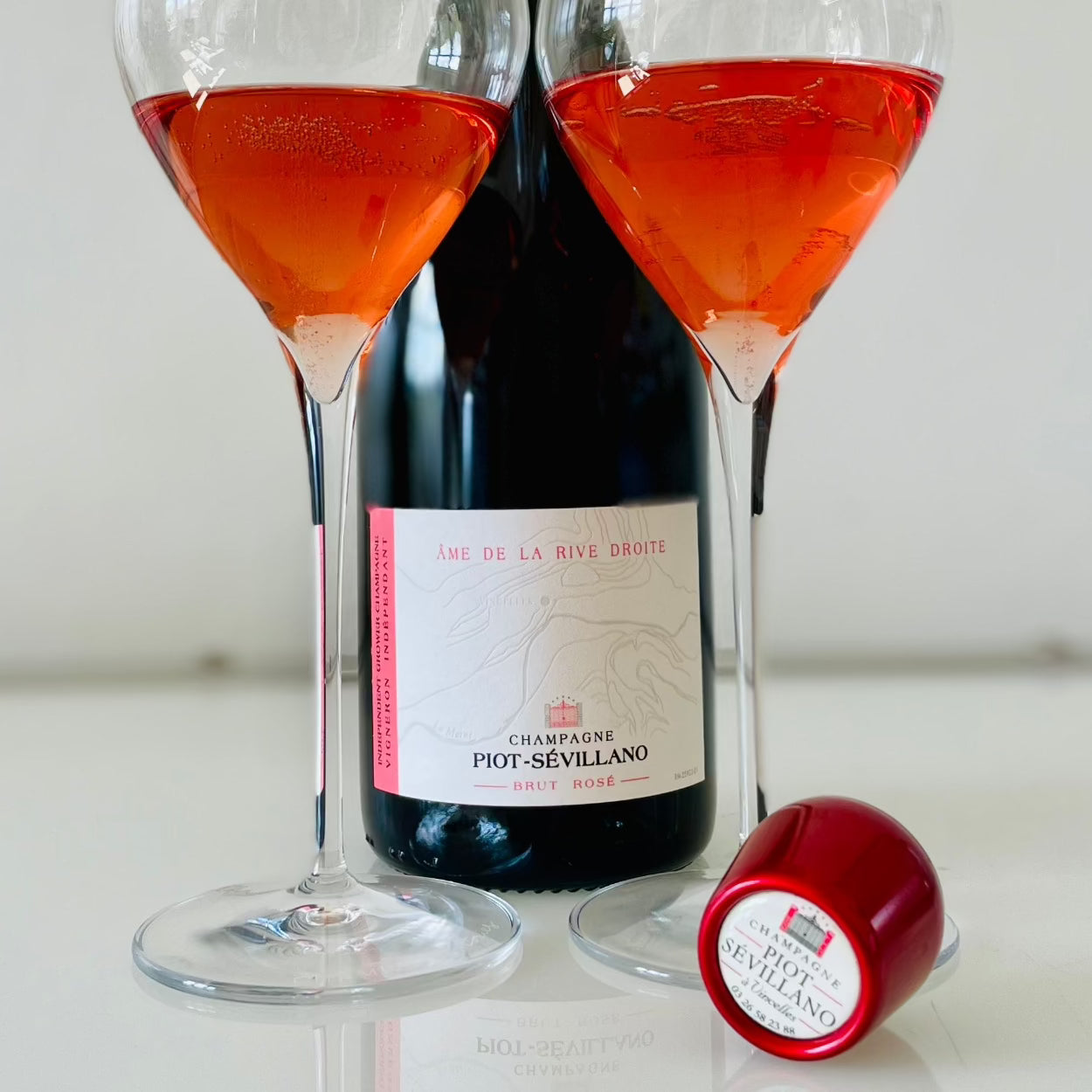 2 champagneglas med rosé champagne på hvidt glasbord, champagneflaske står imellem glassene, rød champagnestopper med logo " Piot-Sévillano", Champagne Piot-Sévillano, Âme de la Rive Droite, Brut Rosé