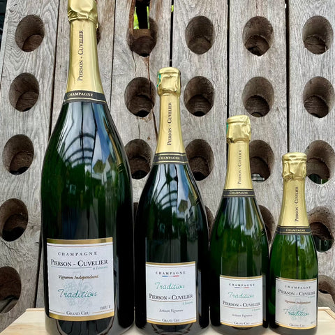 4 flasker champagne fra Champagne Pierson-Cuvelier, Tradition, Grand Cru, 4 forskellige størrelser flaske, 1/2 flaske 0,37 liter, normal flaske 0,75 liter, magnum flaske 1,5 liter, jerobaum 3 liter, baggrund er en pupitre, træ plade med huller til champagneflasker